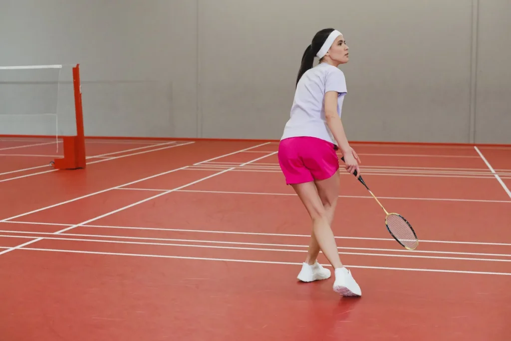 A badminton player in a white top and pink shorts turning on an indoor court, with her racket held low