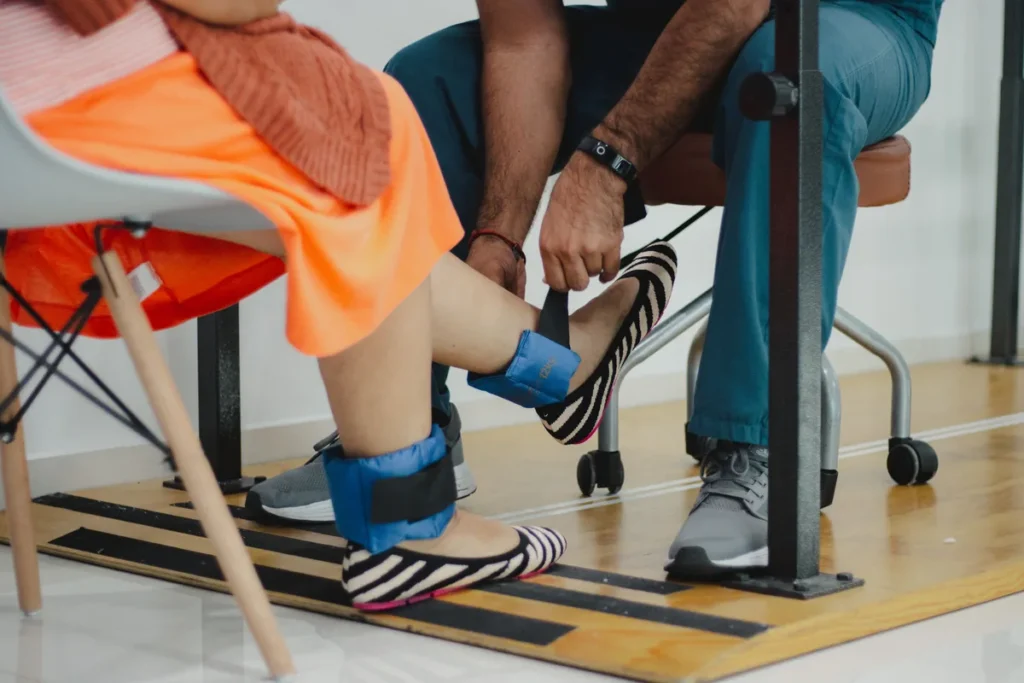 A physiotherapist fastening a weight around a seated patient's ankle, with a second weight already on her other ankle