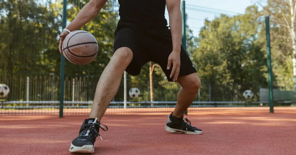 A basketball player in black dribbling low on an outdoor court, knees bent and weight shifted onto one leg