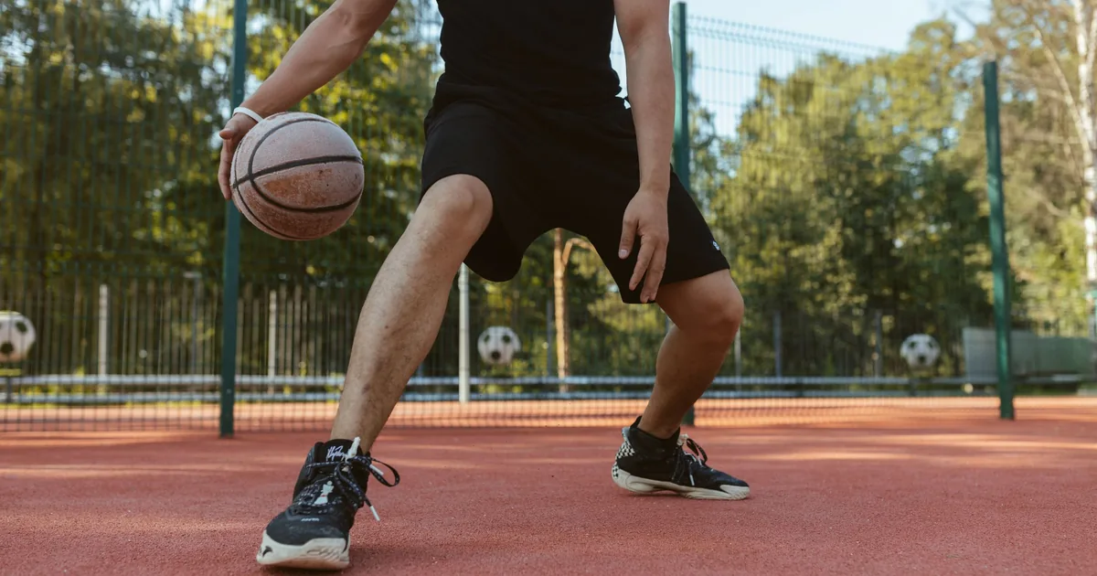 A basketball player in black dribbling low on an outdoor court, knees bent and weight shifted onto one leg