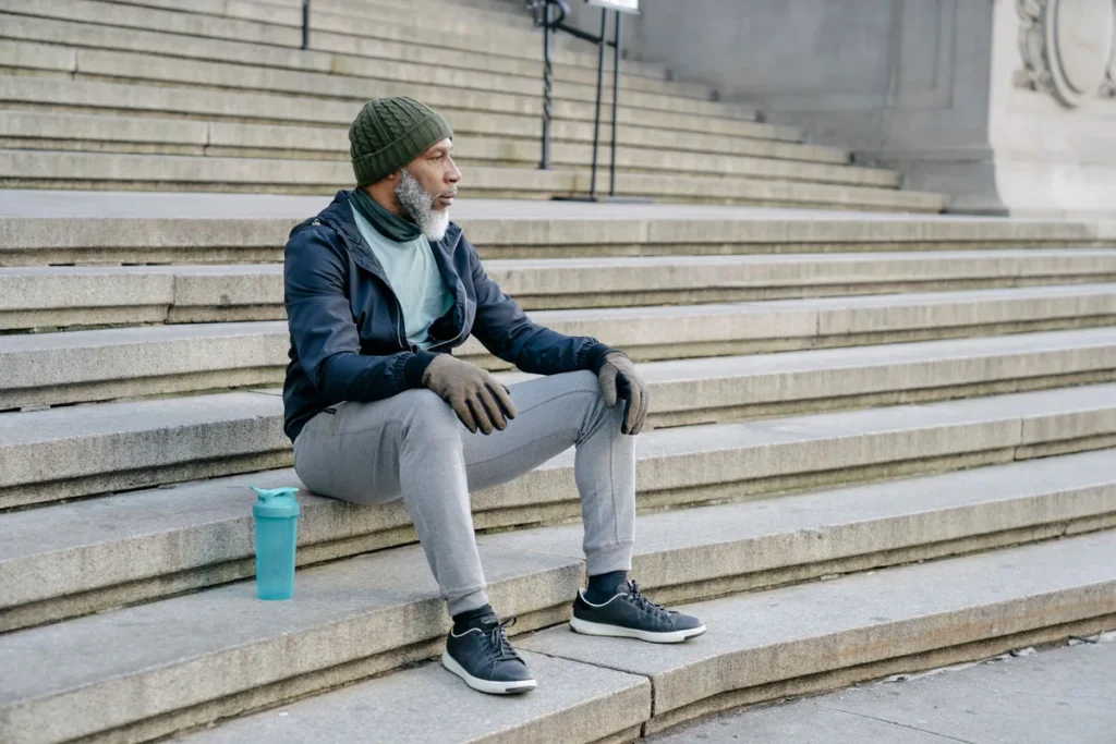 An older man in a knitted hat and sportswear sitting on wide stone steps, resting with a water bottle beside him